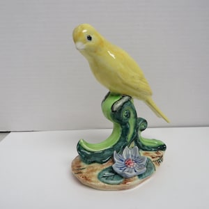 May include: A ceramic figurine of a yellow bird perched on a green, leafy branch. The base features a blue and pink flower with green leaves. The bird has a glossy finish and is set against a neutral background.