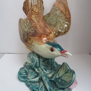 May include: A ceramic bird figurine with brown, green, and blue paint, perched on a green leaf base. The bird has its wings spread and its beak open.