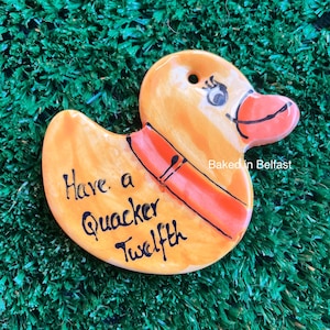 May include: Yellow ceramic duck ornament with a red and orange scarf. The duck has a black eye and a red beak. The ornament says "Have a Quacker Twelfth" and "Baked in Belfast".