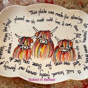 May include: A white ceramic serving plate with scalloped edges featuring a hand-painted illustration of three Highland cows in vibrant rainbow colours. The plate includes black handwritten text circling the cows and the text Baked in Belfast.