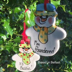 May include: Two hand-painted ceramic snowman ornaments with red and green accents. The larger snowman has a blue hat and a yellow scarf with green polka dots. It says "I'm foundered" and "Baked in Belfast". The smaller snowman has a red hat and a green scarf with red polka dots. It says "It's Baltic!"