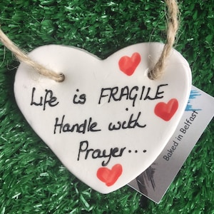 Puede incluir: Adorno de cerámica blanca en forma de corazón con el texto "Life is FRAGILE Handle with Prayer..." escrito en cursiva negra. Hay cuatro corazones rojos pintados en el adorno. El adorno está colgado de una cuerda de yute.