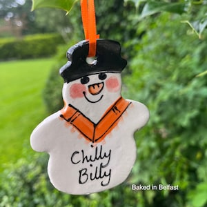 Può includere: Un ornamento di ceramica bianca a forma di pupazzo di neve con un cappello nero, una sciarpa arancione e un viso dipinto. Il pupazzo di neve ha un naso di carota e un sorriso. L'ornamento è decorato con le parole "Chilly Billy" e "Baked in Belfast".
