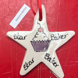 May include: A white ceramic star-shaped ornament with a red ribbon. The ornament features a cupcake illustration and the words "Star Baker" in black. A tag that says "Baked in Belfast" is attached.