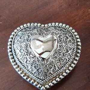 May include: A silver heart-shaped trinket box with an ornate design and a raised heart in the center.