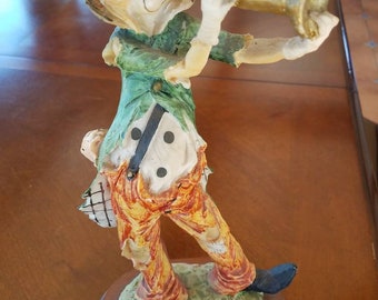 May include: A ceramic clown figurine playing a trumpet. The clown is wearing a green jacket, orange pants, and a black hat. The figurine is standing on a wooden base.