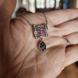 May include: Silver necklace with a pendant featuring a rectangular top with pink gemstones and clear stones, and a teardrop-shaped bottom with black gemstones. The necklace is held in a hand.