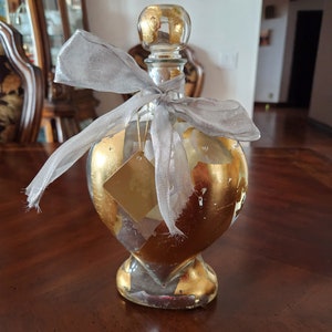 May include: A clear glass bottle with a heart-shaped body and a gold leaf design. The bottle is tied with a gray ribbon and has a gold leaf stopper.