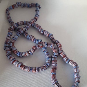 May include: A long strand of blue and red striped glass beads. The beads are cylindrical and have a white stripe running down the center.