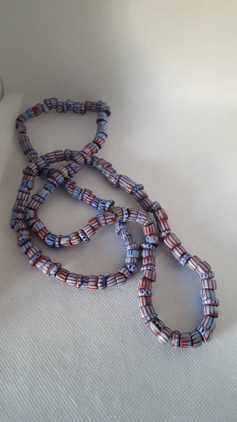 May include: A long strand of blue and red striped glass beads. The beads are cylindrical and have a white stripe running down the center.