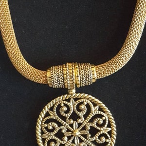 May include: A gold necklace with a round, intricate pendant featuring a floral design. The necklace is made of a woven chain.