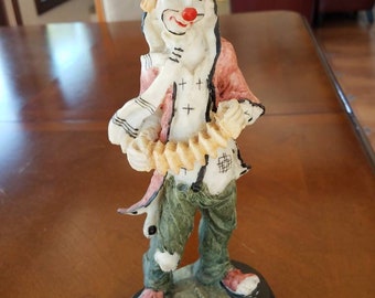 May include: A figurine of a clown with a red nose, wearing a black hat, a red and white shirt, and green pants. The clown is holding a musical instrument and is standing on a black base.