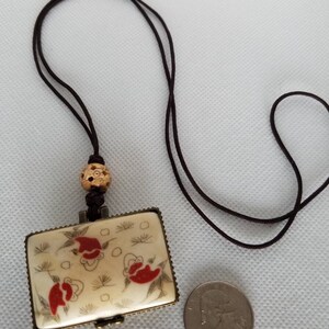 May include: A small, rectangular, white box with a floral design and red accents hangs from a brown cord. The box has a hinged lid and is attached to a brown cord with a wooden bead. A US quarter is shown for scale.