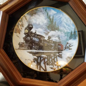May include: A framed porcelain plate with a painting of a steam train traveling through a snowy mountain landscape. The train is numbered "15" and the plate is signed "Tom Yeatts".