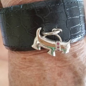 May include: A black leather bracelet with a silver dog charm. The dog charm has a red stone on its collar.