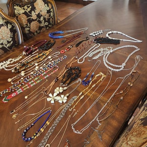 May include: A collection of 20 necklaces with various styles and materials, including beads, shells, and metal chains. The necklaces are laid out on a wooden table.