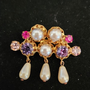 May include: A gold-tone brooch with a cluster of purple, pink, and white stones. The brooch has three dangling pearls.