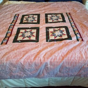 May include: A red and white checkered quilt with four green and white star-shaped patches. The quilt is on a bed with a white headboard and a white and blue pillow.