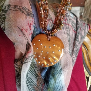 May include: A multi-strand necklace with brown and silver beads and a large heart-shaped pendant. The pendant is made of a copper-colored material with a speckled pattern and is accented with small silver studs.
