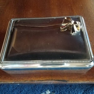 May include: A silver rectangular box with a decorative bow on the lid. The box has a beaded border around the edge.
