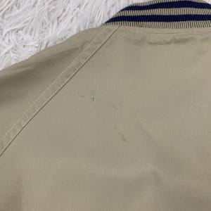 K&ouml;nnte beinhalten: Nahaufnahme einer beigefarbenen Jacke mit einem marineblau-beige gestreiften Kragen. Die Jacke hat ein schlichtes Design mit sichtbaren N&auml;hten entlang der N&auml;hte. Der Stoff scheint ein leichtes Material zu sein.