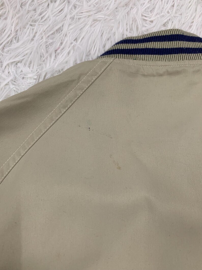 K&ouml;nnte beinhalten: Nahaufnahme einer beigefarbenen Jacke mit einem marineblau-beige gestreiften Kragen. Die Jacke hat ein schlichtes Design mit sichtbaren N&auml;hten entlang der N&auml;hte. Der Stoff scheint ein leichtes Material zu sein.