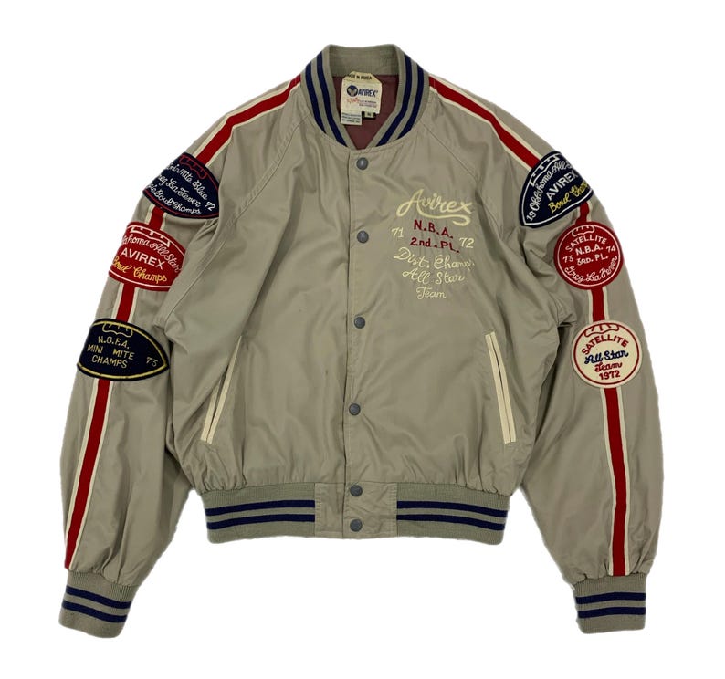K&ouml;nnte beinhalten: Beige Bomberjacke mit roten und wei&szlig;en Streifen auf den &Auml;rmeln. Die Jacke hat mehrere Aufn&auml;her mit Text, darunter "Avirex", "N.B.A." und "Satellite All Star Team". Sie hat eine Knopfleiste und einen gerippten Kragen und B&uuml;ndchen.