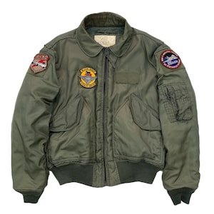 May include: Olive green bomber jacket with various patches. The jacket has a zip-up front, two front pockets, and ribbed cuffs and hem. Patches include text and graphic designs. A-10 and Black Sheep Stealth Driven patches are visible.