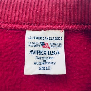 May include: Close-up of a red sweatshirt collar and tag. The tag reads "ALL-AMERICAN CLASSICS" and "AVIREX U.S.A." with a small American flag graphic. The tag also indicates "Certificate of Authenticity" and "Small" size.