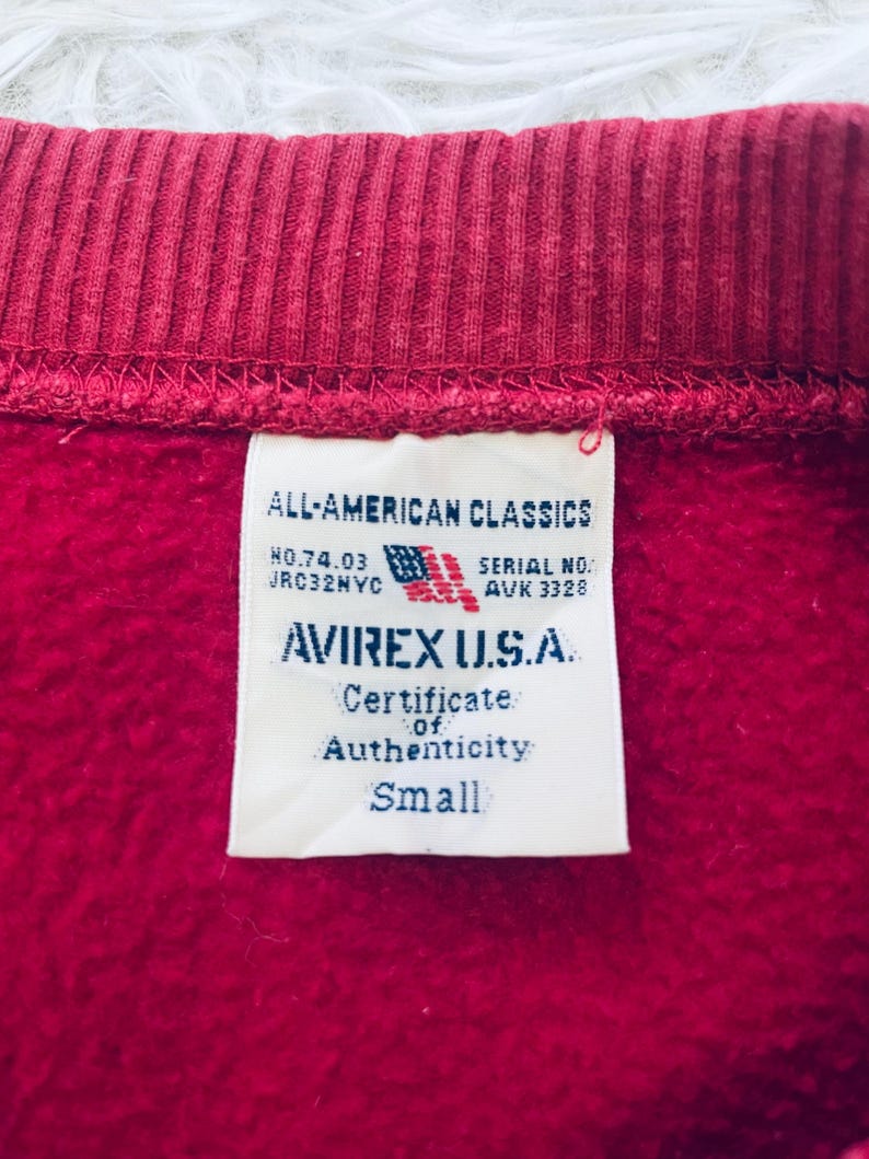 May include: Close-up of a red sweatshirt collar and tag. The tag reads "ALL-AMERICAN CLASSICS" and "AVIREX U.S.A." with a small American flag graphic. The tag also indicates "Certificate of Authenticity" and "Small" size.