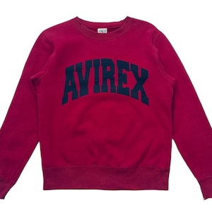 May include: A crimson sweatshirt with the word "AVIREX" arched across the chest in navy blue block letters. The long-sleeved crew neck top has a small tag at the neckline.