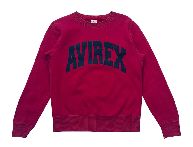 May include: A crimson sweatshirt with the word "AVIREX" arched across the chest in navy blue block letters. The long-sleeved crew neck top has a small tag at the neckline.