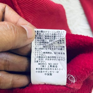 May include: Close-up of a red fabric with a white tag attached. The tag has Japanese text and is partially visible. The fabric appears to be a piece of clothing, possibly a sweatshirt or jacket.