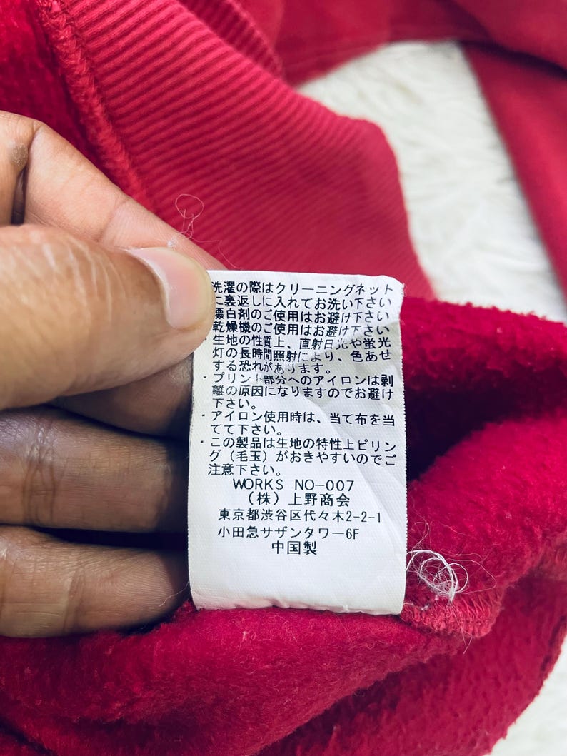 May include: Close-up of a red fabric with a white tag attached. The tag has Japanese text and is partially visible. The fabric appears to be a piece of clothing, possibly a sweatshirt or jacket.