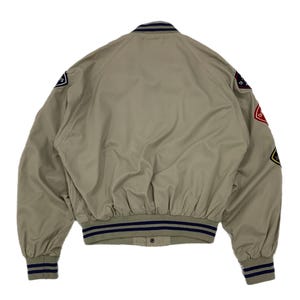 K&ouml;nnte beinhalten: Beige Bomberjacke mit marineblauen und wei&szlig;en Streifen an den B&uuml;ndchen, am Kragen und am Bund. Die Jacke hat Aufn&auml;her auf den &Auml;rmeln mit verschiedenen Designs und Texten. Das Material ist glatt und leicht.