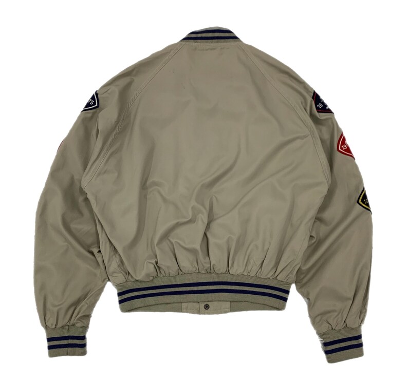 K&ouml;nnte beinhalten: Beige Bomberjacke mit marineblauen und wei&szlig;en Streifen an den B&uuml;ndchen, am Kragen und am Bund. Die Jacke hat Aufn&auml;her auf den &Auml;rmeln mit verschiedenen Designs und Texten. Das Material ist glatt und leicht.