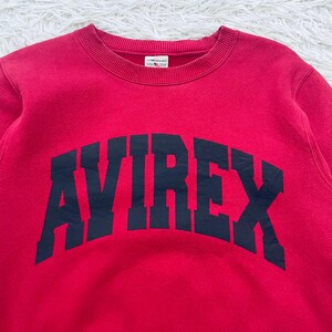 May include: A red crewneck sweatshirt with the word "AVIREX" arched across the chest in navy blue block letters. The sweatshirt has a ribbed neckline and cuffs. The fabric appears to be a soft, comfortable material.