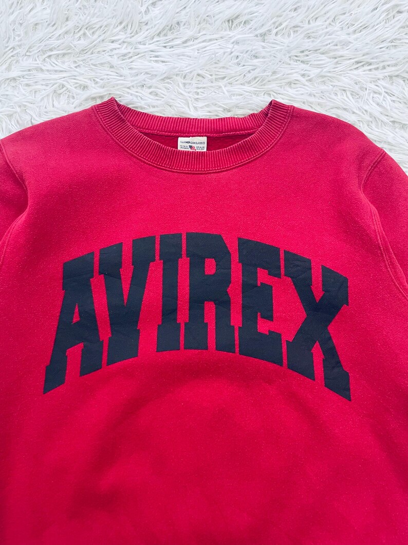 May include: A red crewneck sweatshirt with the word "AVIREX" arched across the chest in navy blue block letters. The sweatshirt has a ribbed neckline and cuffs. The fabric appears to be a soft, comfortable material.