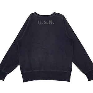Può includere: Una felpa blu navy con le lettere "U.S.N." stampate sulla parte superiore della schiena. La parte superiore a maniche lunghe ha polsini a coste e girocollo. Il tessuto sembra essere un materiale morbido e usurato.