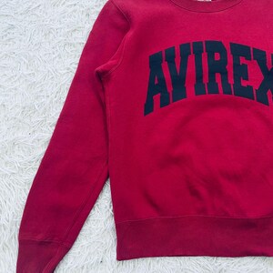 May include: A red sweatshirt with the word "AVIREX" in large, navy blue block letters across the chest. The sweatshirt has a crew neck and ribbed cuffs and hem. The fabric appears to be a soft, comfortable material.