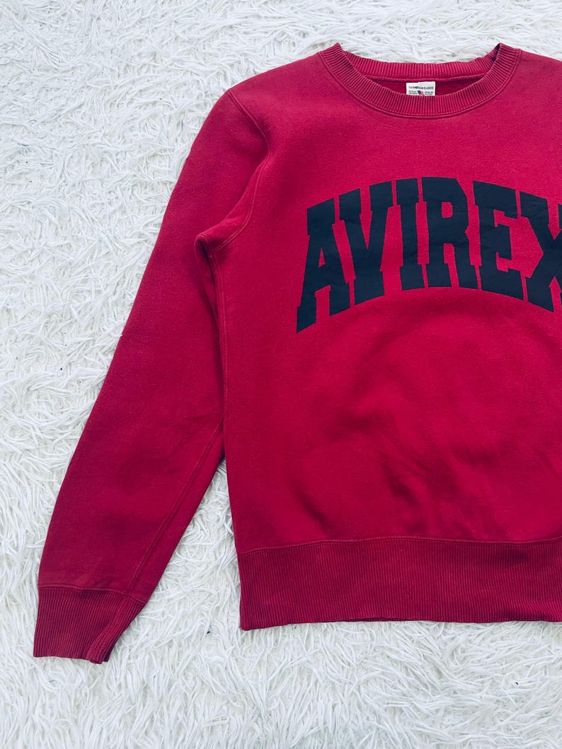 May include: A red sweatshirt with the word "AVIREX" in large, navy blue block letters across the chest. The sweatshirt has a crew neck and ribbed cuffs and hem. The fabric appears to be a soft, comfortable material.