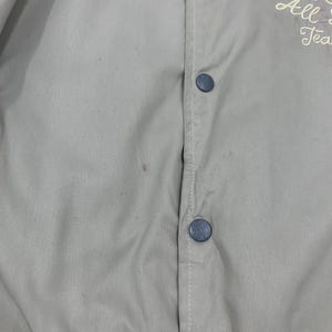 K&ouml;nnte beinhalten: Hellbeige Jacke mit Knopfleiste und blauen Kn&ouml;pfen. Wei&szlig;e N&auml;hte verlaufen entlang der Vorderseite. Auf der Jacke ist Text in Rot und Wei&szlig; gestickt. Das Kleidungsst&uuml;ck scheint Vintage zu sein.