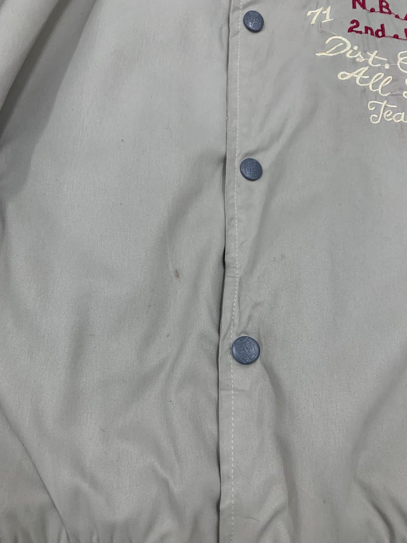 K&ouml;nnte beinhalten: Hellbeige Jacke mit Knopfleiste und blauen Kn&ouml;pfen. Wei&szlig;e N&auml;hte verlaufen entlang der Vorderseite. Auf der Jacke ist Text in Rot und Wei&szlig; gestickt. Das Kleidungsst&uuml;ck scheint Vintage zu sein.