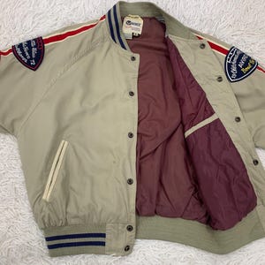 K&ouml;nnte beinhalten: Beigefarbene Bomberjacke mit roten und wei&szlig;en Streifen auf den &Auml;rmeln. Die Jacke hat Aufn&auml;her auf den &Auml;rmeln mit Text und ein weinrotes Innenfutter. Die Jacke hat eine Knopfleiste und zwei Vordertaschen.