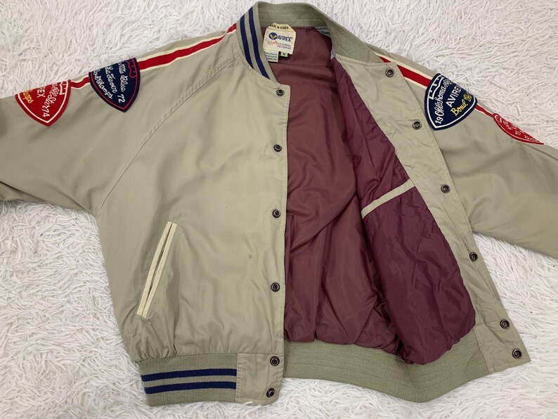 K&ouml;nnte beinhalten: Beigefarbene Bomberjacke mit roten und wei&szlig;en Streifen auf den &Auml;rmeln. Die Jacke hat Aufn&auml;her auf den &Auml;rmeln mit Text und ein weinrotes Innenfutter. Die Jacke hat eine Knopfleiste und zwei Vordertaschen.
