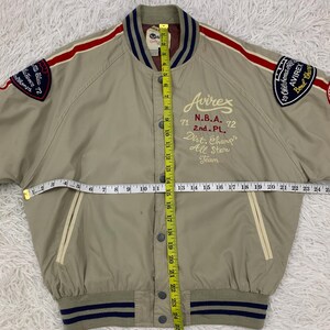 K&ouml;nnte beinhalten: Beige Bomberjacke mit roten und wei&szlig;en Streifen auf den &Auml;rmeln. Mit Aufn&auml;hern mit Text und dem Avirex-Logo. Die Jacke hat eine Knopfleiste und einen gestreiften Kragen und B&uuml;ndchen. Die Jacke ist ca. 63.5 cm lang.