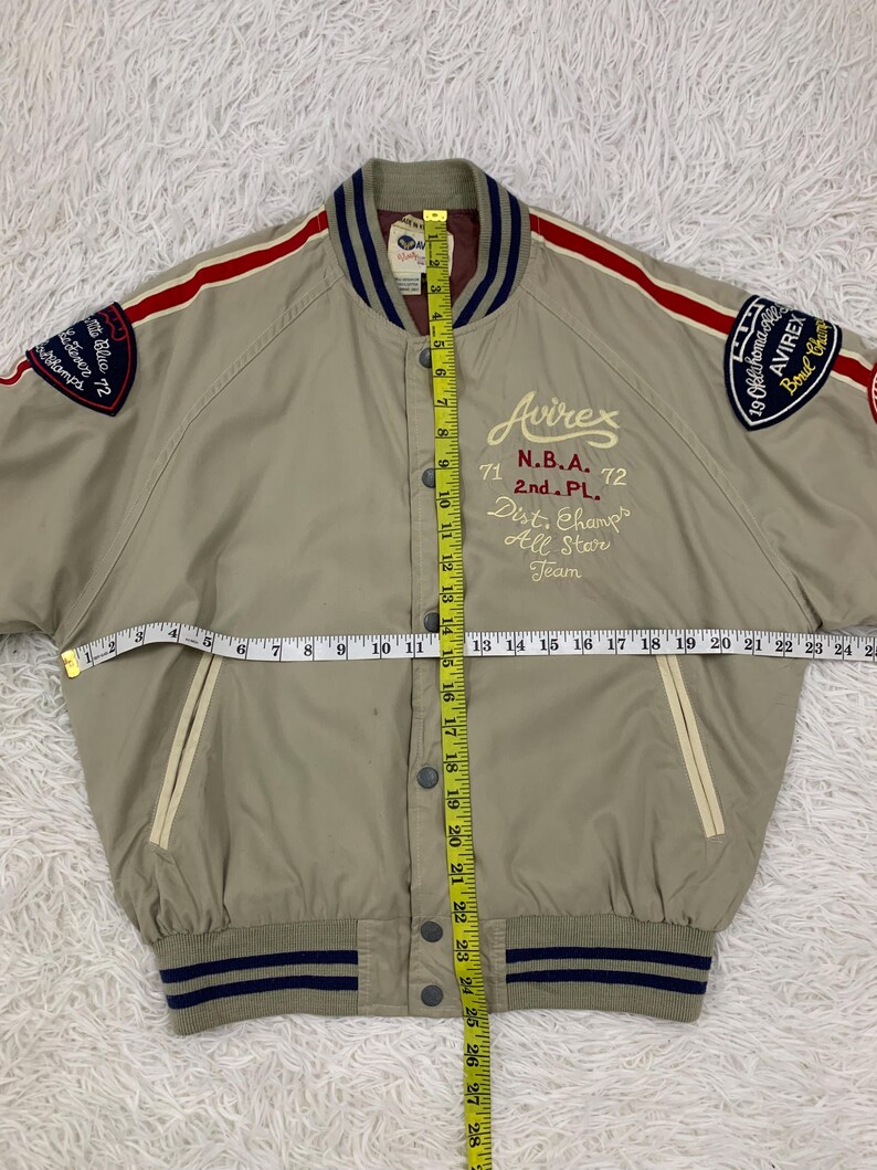 K&ouml;nnte beinhalten: Beige Bomberjacke mit roten und wei&szlig;en Streifen auf den &Auml;rmeln. Mit Aufn&auml;hern mit Text und dem Avirex-Logo. Die Jacke hat eine Knopfleiste und einen gestreiften Kragen und B&uuml;ndchen. Die Jacke ist ca. 63.5 cm lang.