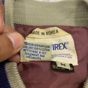 K&ouml;nnte beinhalten: Nahaufnahme eines Vintage-Bomberjacken-Etiketts. Das Etikett lautet "MADE IN KOREA" und enth&auml;lt Angaben zur Stoffzusammensetzung und Pflegehinweise. Das Innere der Jacke ist bordeauxfarben, mit einem beigen Kragen und blauen Streifen.