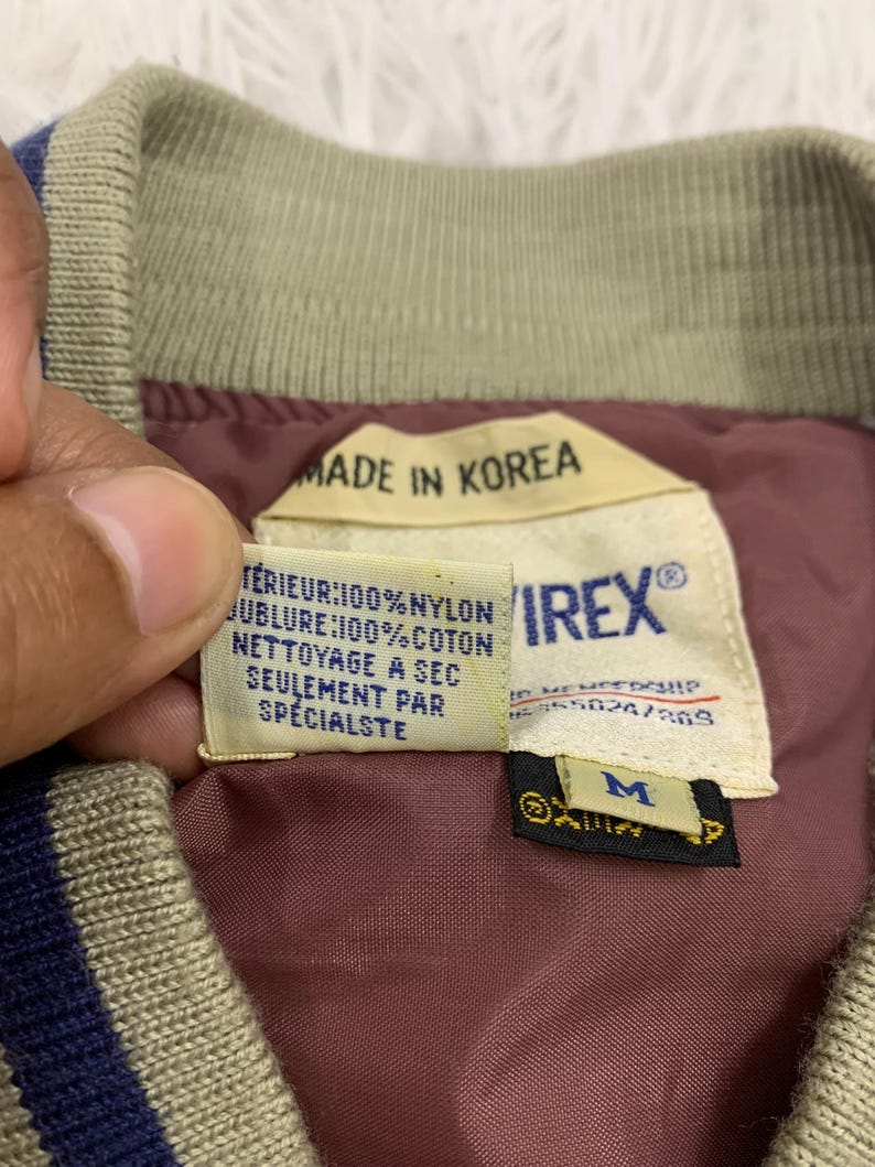 K&ouml;nnte beinhalten: Nahaufnahme eines Vintage-Bomberjacken-Etiketts. Das Etikett lautet "MADE IN KOREA" und enth&auml;lt Angaben zur Stoffzusammensetzung und Pflegehinweise. Das Innere der Jacke ist bordeauxfarben, mit einem beigen Kragen und blauen Streifen.