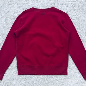 May include: A solid crimson sweatshirt with a crew neck and long sleeves. The garment has ribbed cuffs and a ribbed hem. A small logo is visible on the upper back. The sweatshirt is made of a soft, comfortable material.